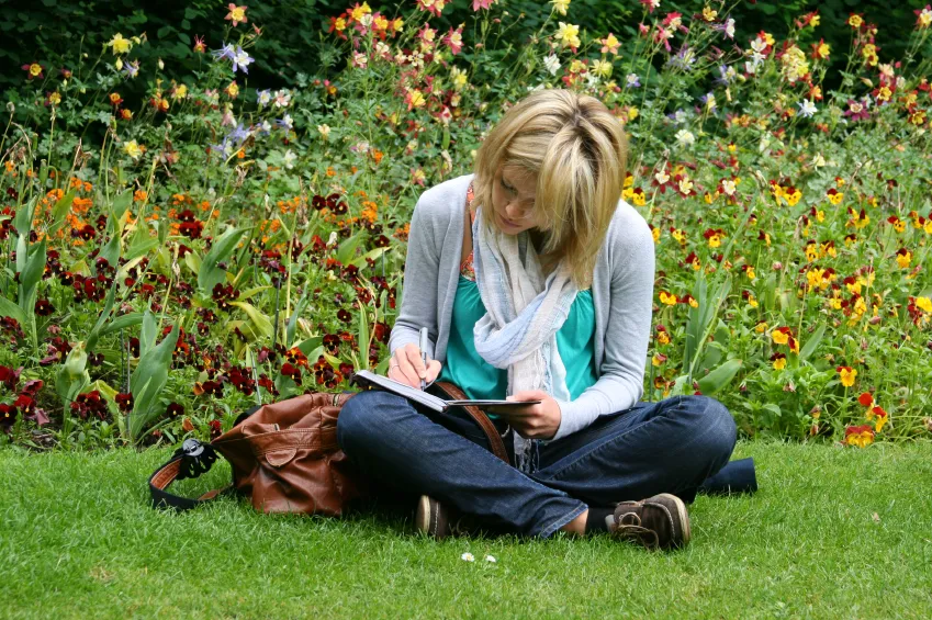 A woman sits in the grass in front of a patch of colorful wildflowers. She sits cross legged, writing in a journal in her lap.