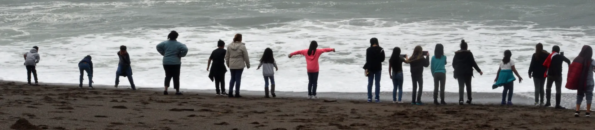 Children and some adults stand on the beach at the water's edge looking at the tides