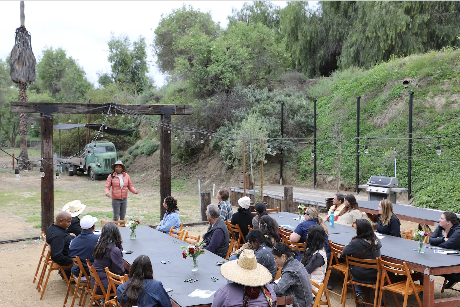 People sit outdoors at long tables listening to a woman speak