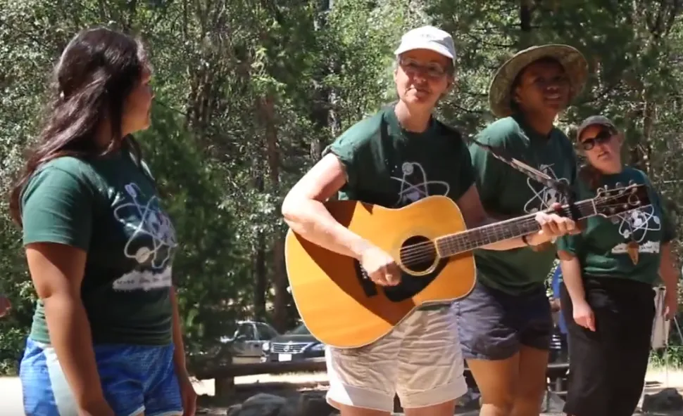 Marianne strums guitar. Three women wearing matching green shirts stand next to her