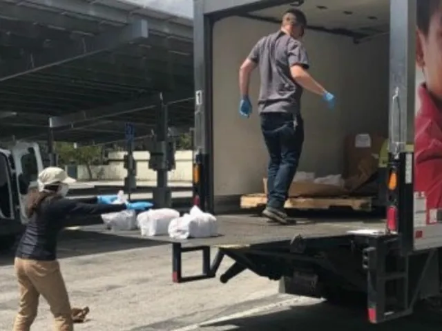 Box truck with man in rubber gloves on the loading dock of truck in a grayish shirt with blue jeans. Woman on lower portion of the truck wearing a mask and rubber gloves, loading food donations.