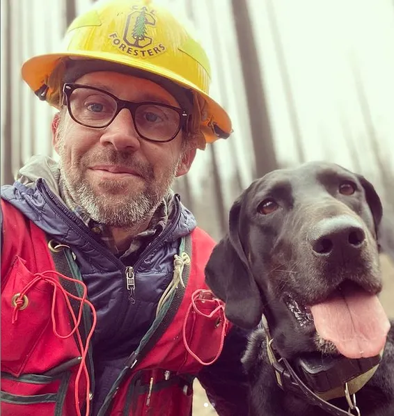 Ryan Tompkins wearing a yellow hard hat and red jacket with his black labrador retriever