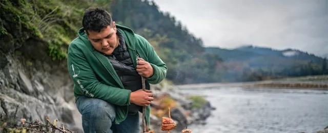 Dark-haired man in a green hoodie works beside a river.