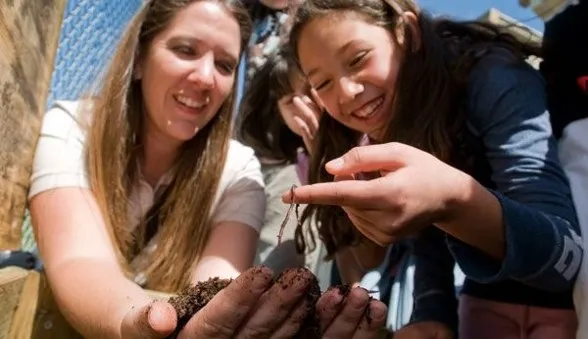Smiling young girl holding a worm on her finger, while adult holds soil in her hands nearby.