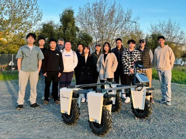 13 people stand around the Amiggie robot