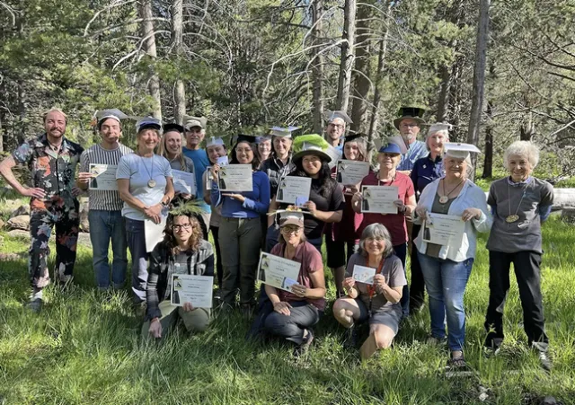 A group of smiling people wearing graduation caps hold up certificates in a grassy, wooded area.