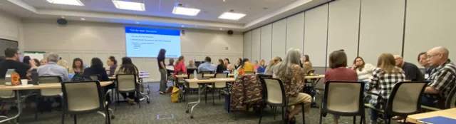 Dozens of people sit at conference room tables. PowerPoint screen reads: