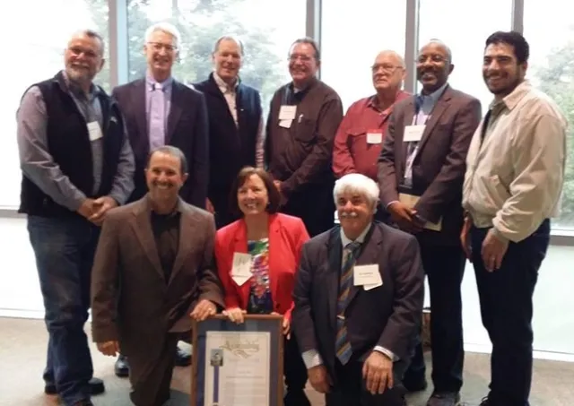 Ten people wearing suits pose with a plaque