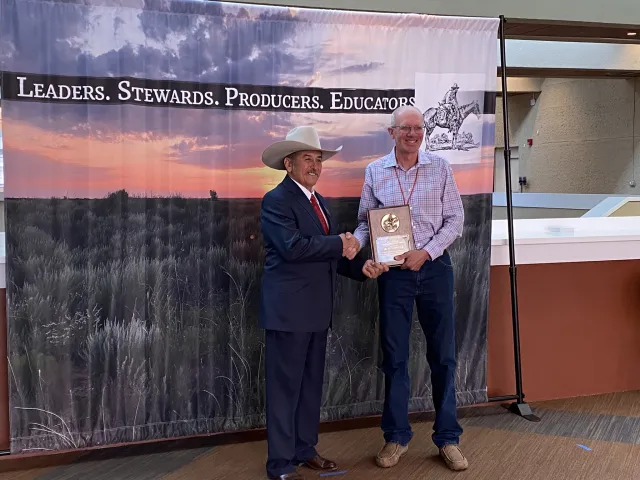 A man wearing a dark suit and beige cowboy hat shakes Ken's hand while handing him an ornate wood plaque.