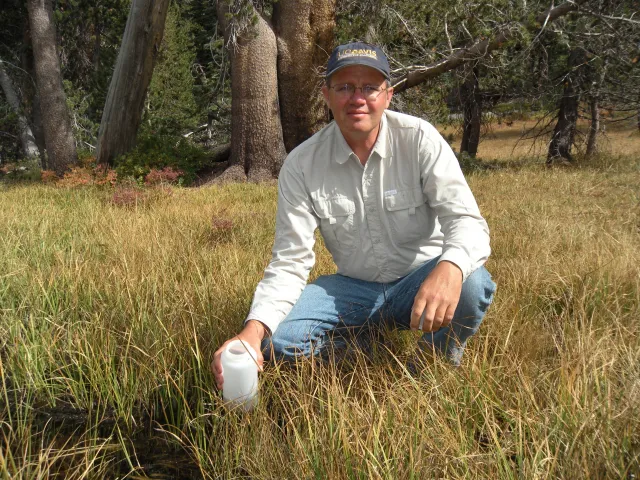 Ken holds a white plastic bottle as he squats in tall grass with trees behind him