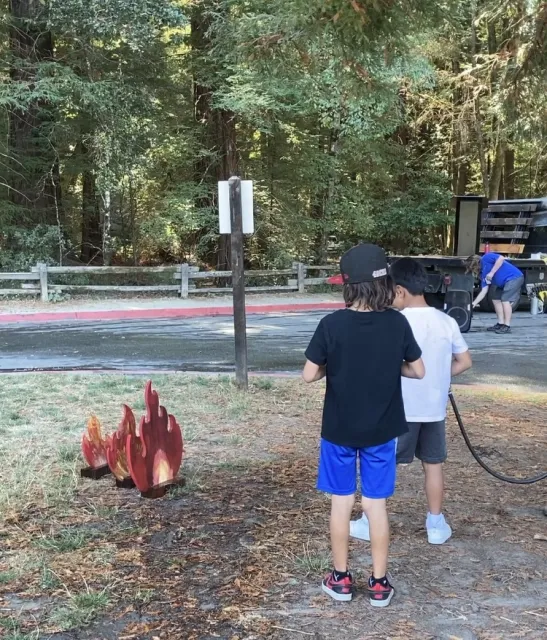 Two boys, one aiming a water hose at wooden cutout of flames