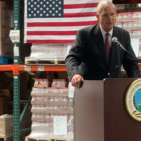 Vilsack stands at lectern with pallets of canned goods on shelves behind him.