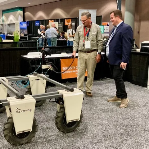 Fennimore controls an automated platform with a handheld device connected to the machine as Youtsey observes. Equipment vendor booths are in the background.