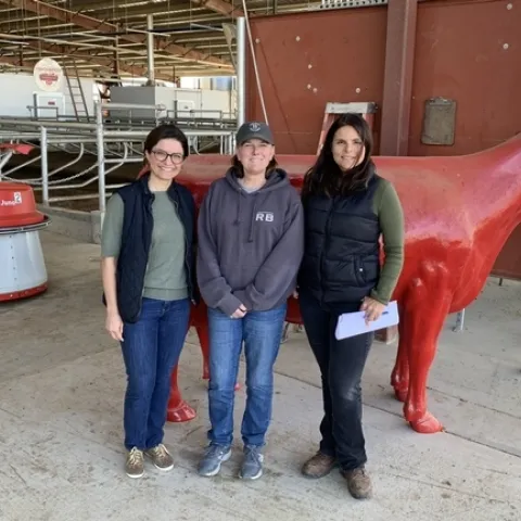De izquierda a derecha: Fernanda Ferreira, ex investigadora de la Facultad de Veterinaria de UC Davis, Shonda Reid, gerente de la lechería Fred Rau y Daniela Bruno, asesora de lecherías de Extensión Cooperativa de UC, quien ha colaborado en estudios sobre lecherías grandes en California que usan los sistemas de ordeño automatizados. A la distancia se observa un robot que empuja alimento hacia las vacas. Fotografía cortesía de Daniela Bruno