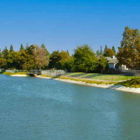 A canal of water next to a fenced area with residential homes and trees.