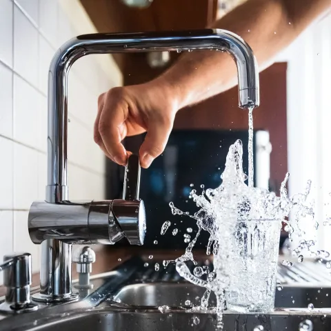 Hand turns on a tap with water splashing out of a glass