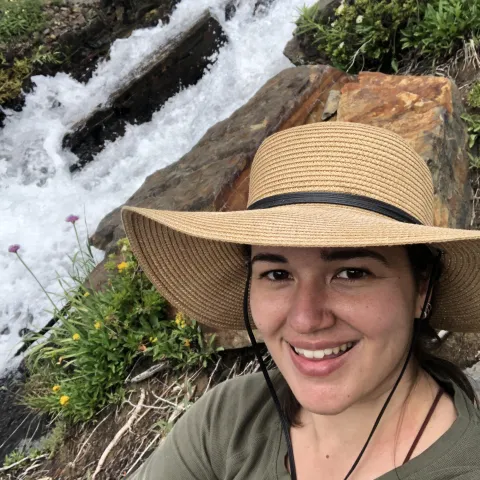 Photo of a woman in a broad sun hat. In the background is a rushing waterfall.