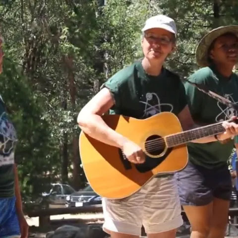 Marianne strums guitar. Three women wearing matching green shirts stand next to her