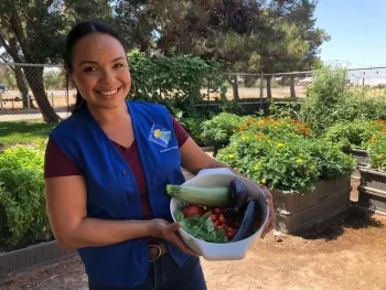 Master Gardener smiling with basket of garden harvest of eggplant, squash, basil, and tomatoes.