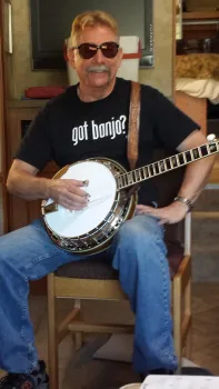 Harper sits in a chair holding a banjo and wearing sunglasses, jeans and a black t-shirt that reads