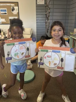 Two school-age girls showing their drawings of a healthy plate of food.