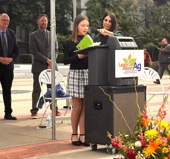 Jayna standing at a podium reading her story at the State Capitol.