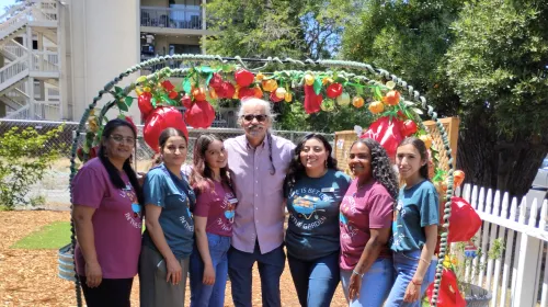 Santos stands beneath an arch decorated with red, yellow and green vegetable ornaments. Three women stand on both sdies of him
