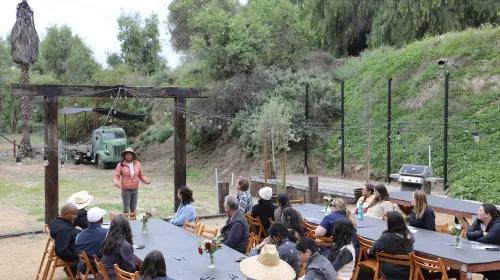 People sit outdoors at long tables listening to a woman speak