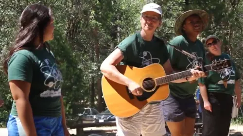 Marianne strums guitar surrounded by 3 other women wearing matching dark green t-shirts